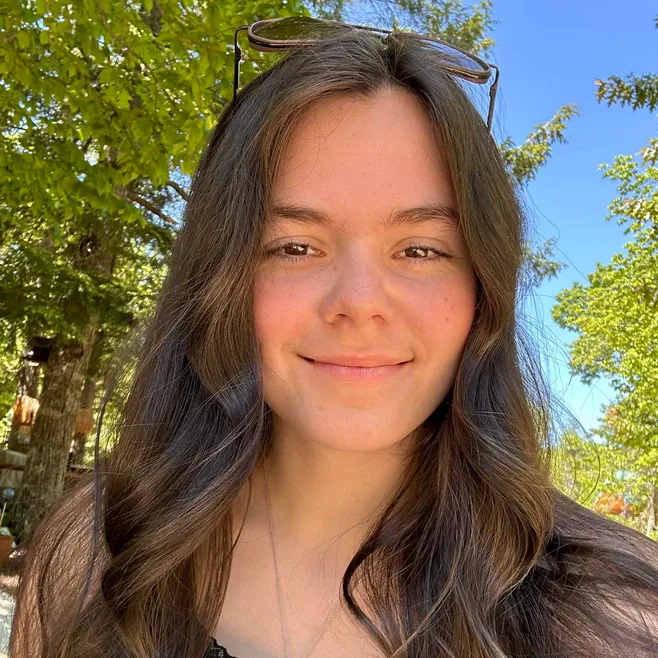 Autumn Mowery, smiling, with sunglasses pushed up into her hair, in front of green trees and a blue sky.