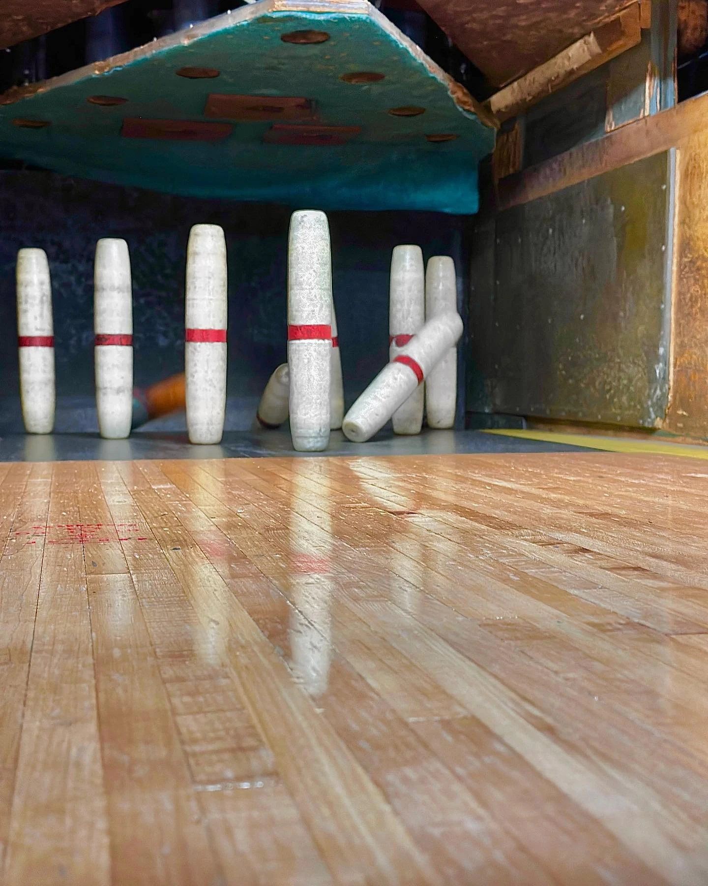Candlepin bowling pins standing at the end of a polished wooden lane, one of them knocked over.