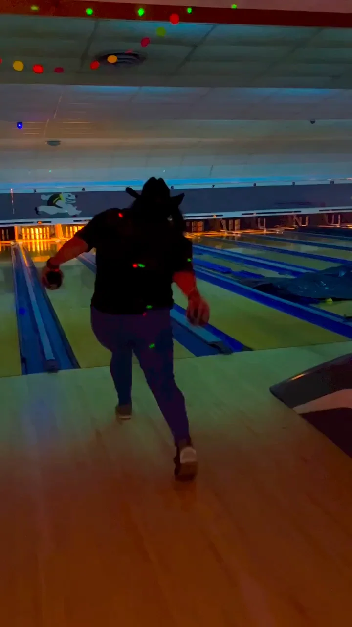 A bowler in a cowboy hat sends a ball down a dimly lit lane.